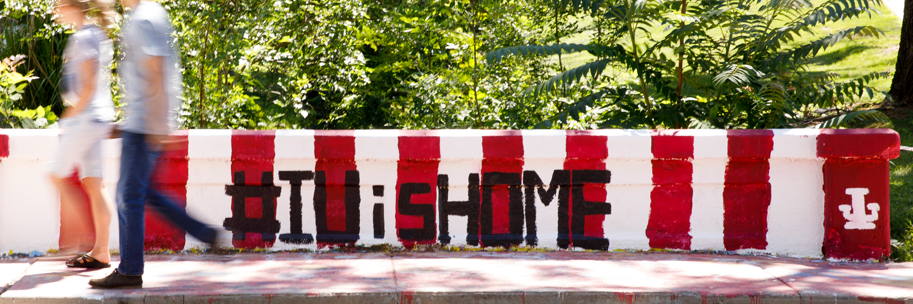 Students walk past a bridge railing painted in red and white stripes and which says #IUisHome.